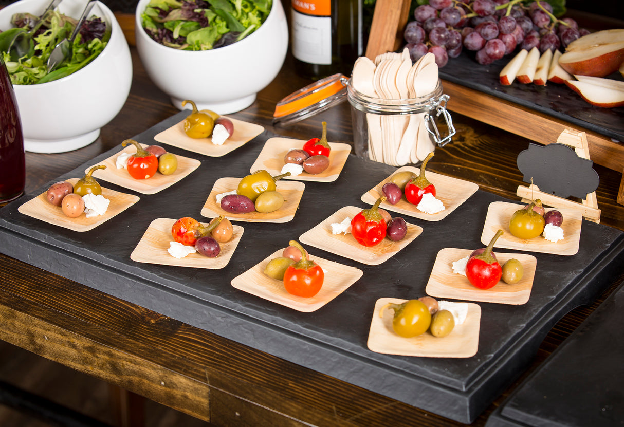 A display of small wooden plates holds stuffed peppers, olives, and cheese cubes on a Frostone Collection Display Tray. In the background are bowls of salad, grapes, sliced pear, and a blank mini chalkboard sign on a buffet table.