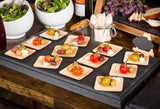 A display of small wooden plates holds stuffed peppers, olives, and cheese cubes on a Frostone Collection Display Tray. In the background are bowls of salad, grapes, sliced pear, and a blank mini chalkboard sign on a buffet table.