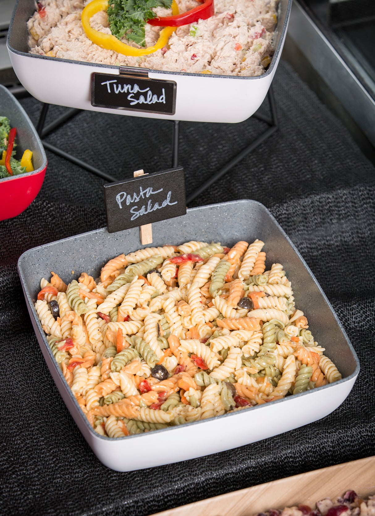A square melamine bowl from the TableCraft Frostone Naturals collection holds tri-color rotini pasta salad with olives and diced vegetables. A small black sign labeled Pasta Salad is placed in the bowl, with Tuna Salad behind it.
