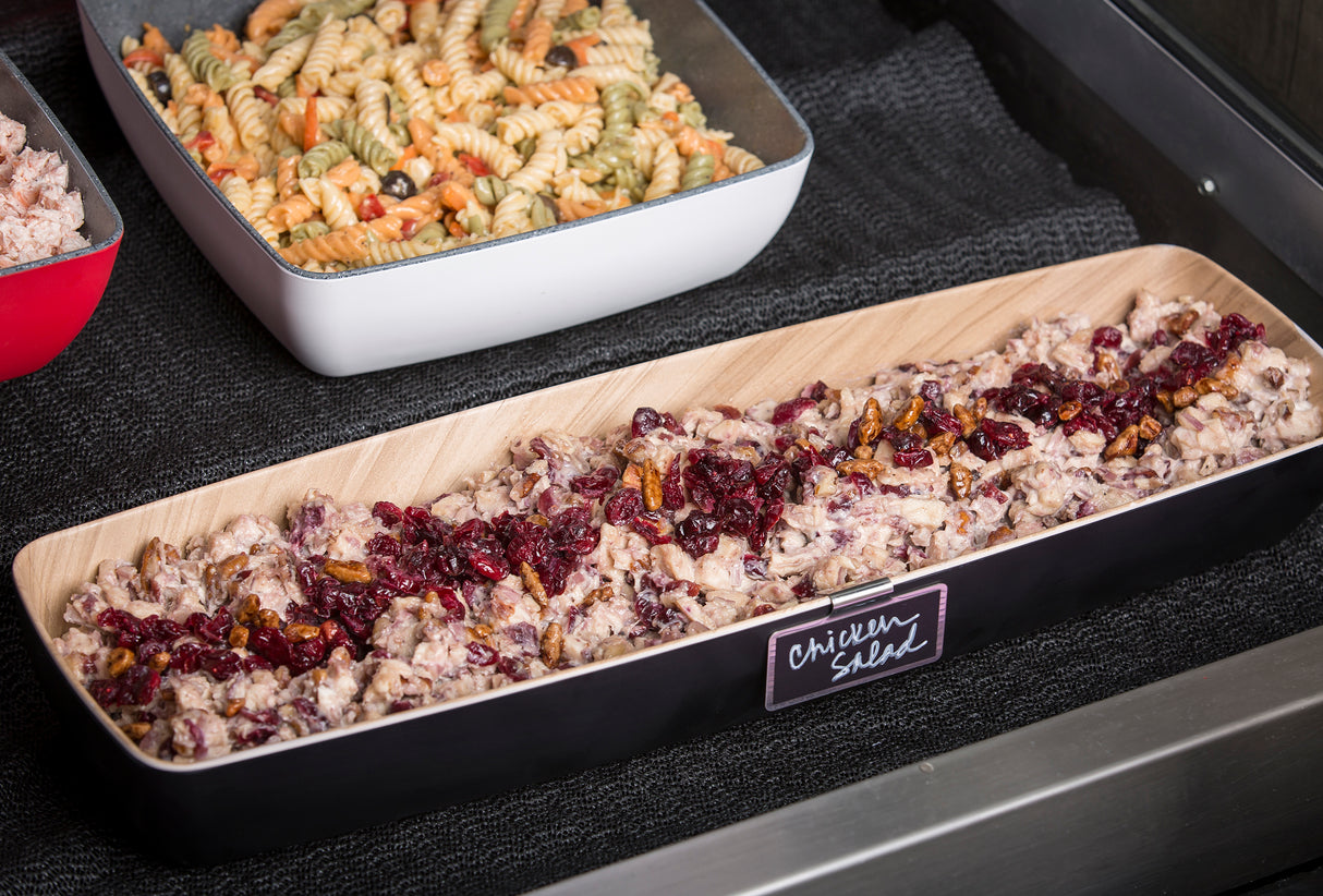 A rectangular wooden tray labeled chicken salad contains a creamy chicken mixture topped with dried cranberries and pecans. Behind it, a TableCraft Products melamine bowl holds tri-color rotini pasta salad, both set on a black cloth surface.
