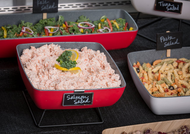 A red Frostone Naturals Bowl filled with salmon salad, garnished with parsley and lemon slices, sits on a black stand. A handwritten label reads Salmon Salad. Nearby are dishes of colorful pasta salad and leafy green salad.