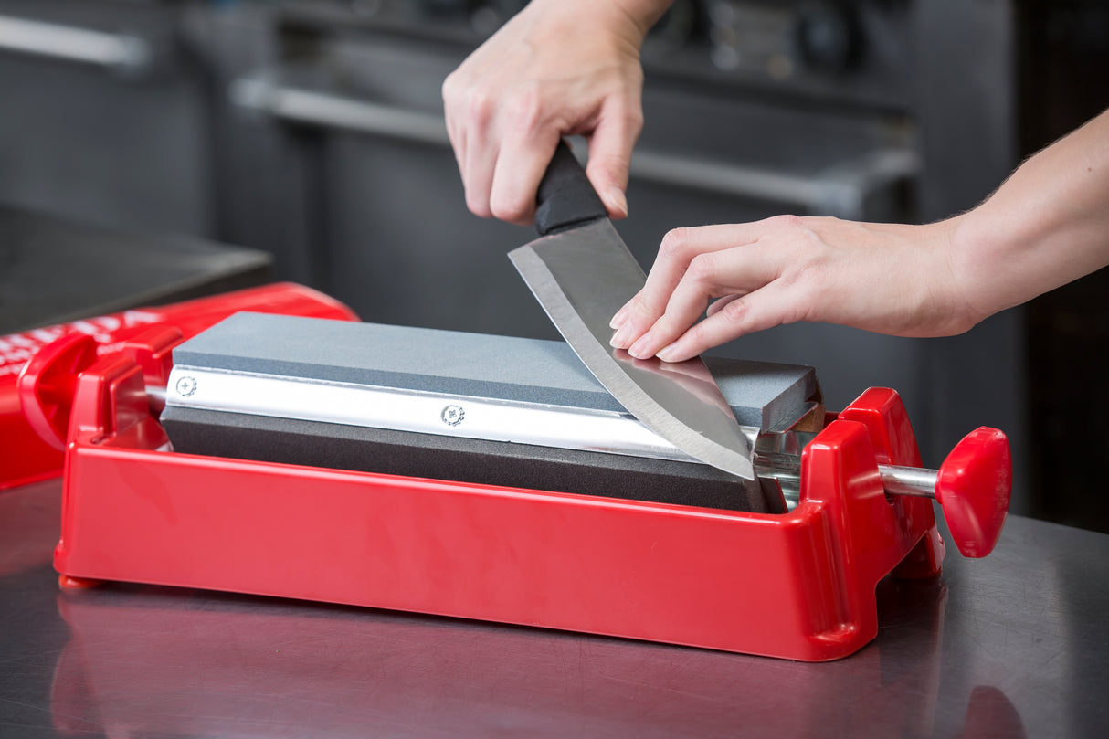 A person sharpens a large kitchen knife on a premium sharpening stone held by a red guide, with both hands visible. The setup, part of a professional knife sharpening system, rests on a metal countertop in a commercial kitchen.