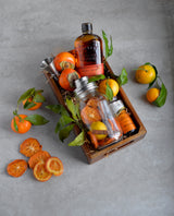 A wooden tray holds a whiskey bottle, Mason Jar Cocktail Shaker with dried fruit slices, stainless steel jigger, and a can. Fresh tangerines and persimmons with green leaves surround the tray on a light gray surface.