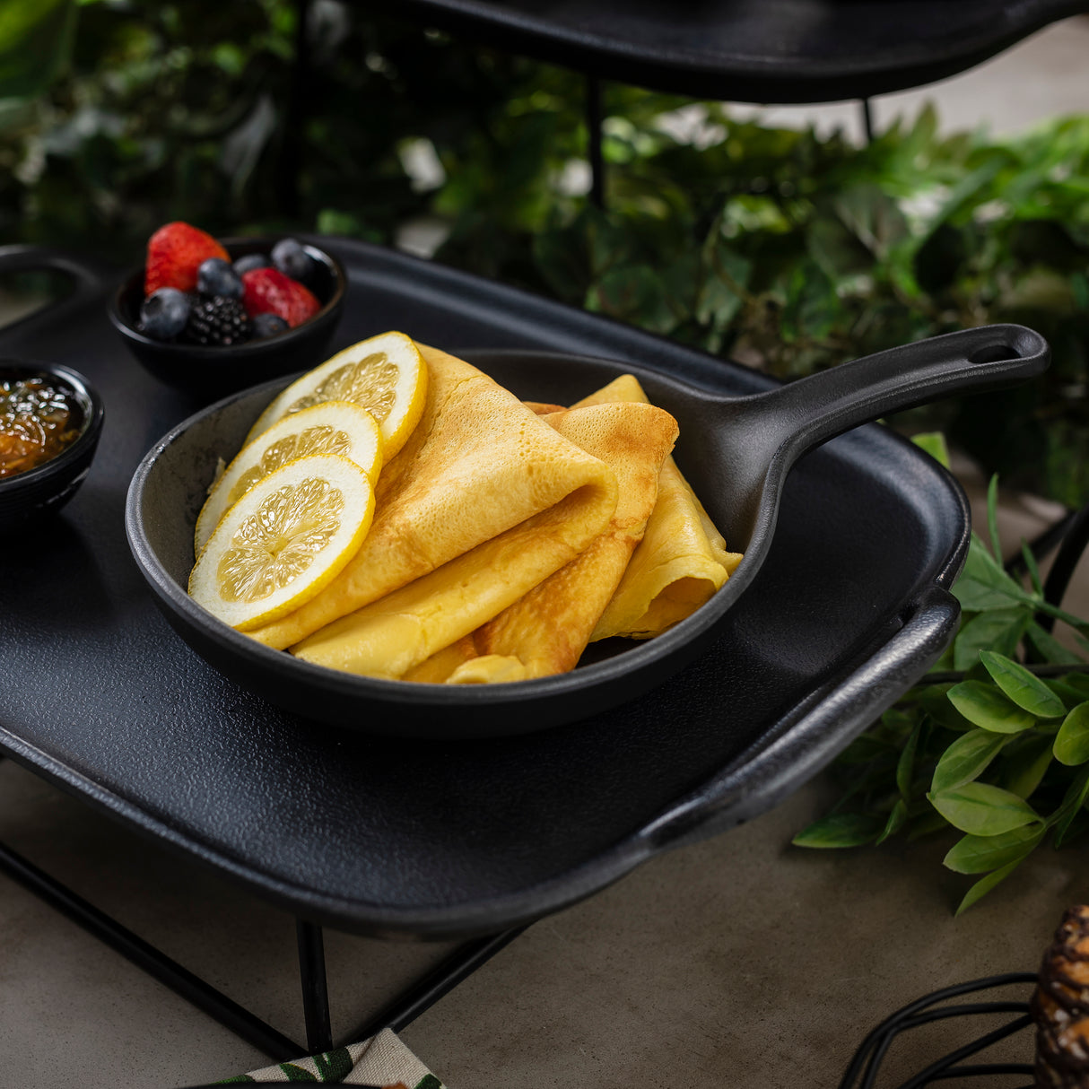 A small black melamine skillet holds folded crepes topped with lemon slices. Next to the skillet are bowls of mixed berries and jam, all set on a black tray with green leaves and plants in the background.