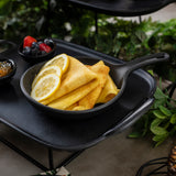 A small black melamine skillet holds folded crepes topped with lemon slices. Next to the skillet are bowls of mixed berries and jam, all set on a black tray with green leaves and plants in the background.