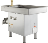 A Hobart Meat Grinder with a 3 HP motor, stainless steel body, and stainless steel pan features a rectangular tray on top, hand crank, metal grinder attachment in front left, sturdy metal legs, and visible safety and brand labels.