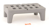 A small, gray, rectangular plastic step stool made from corrosion proof polymer with four wide legs and a slotted top sits on a white surface. In front of the stool, there is a small, orange, bone-shaped object.
