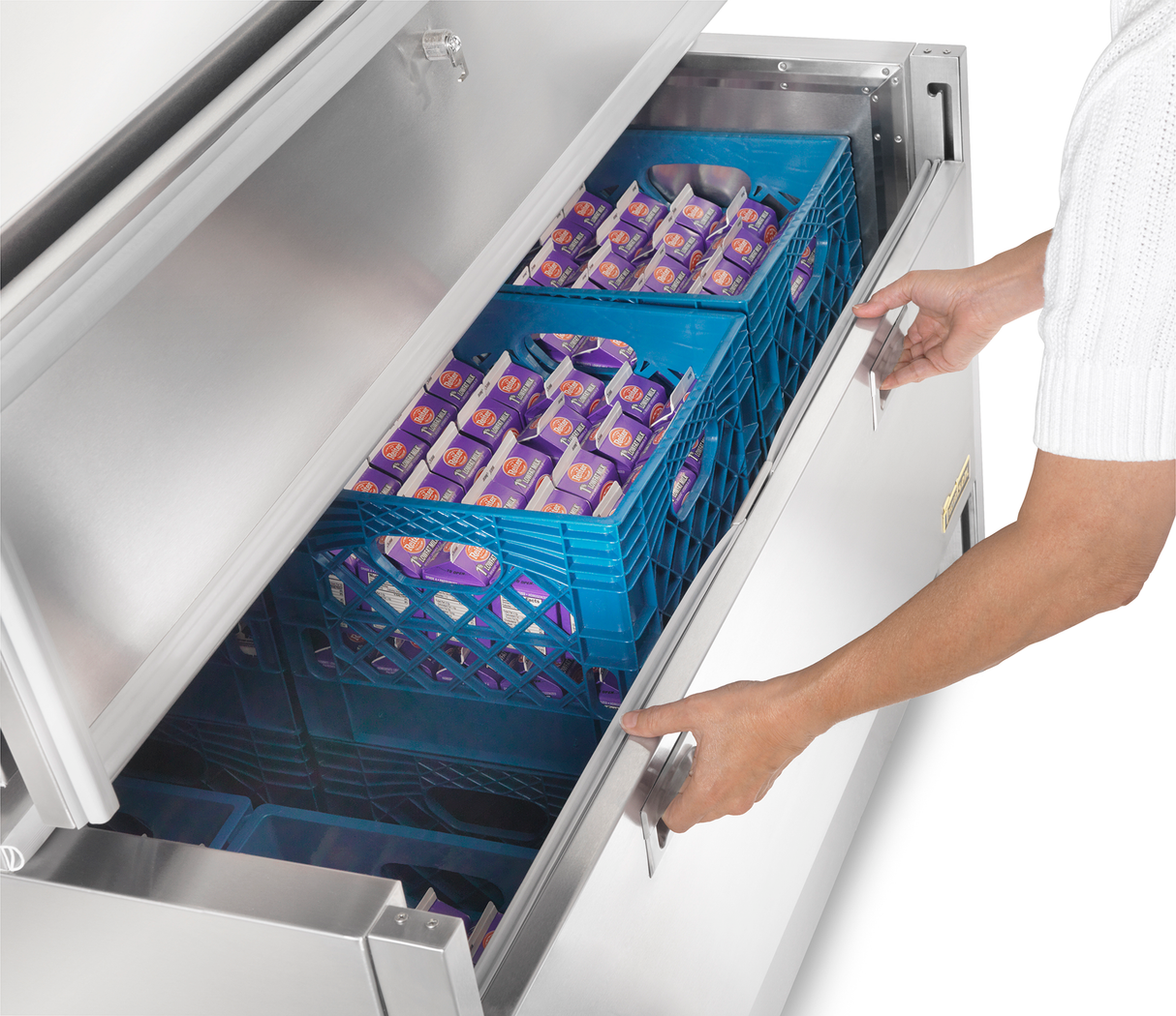 A person in a white shirt opens a large stainless steel milk cooler drawer filled with blue crates holding small purple cartons of milk. The refrigerator appears to be in a commercial or institutional kitchen setting.