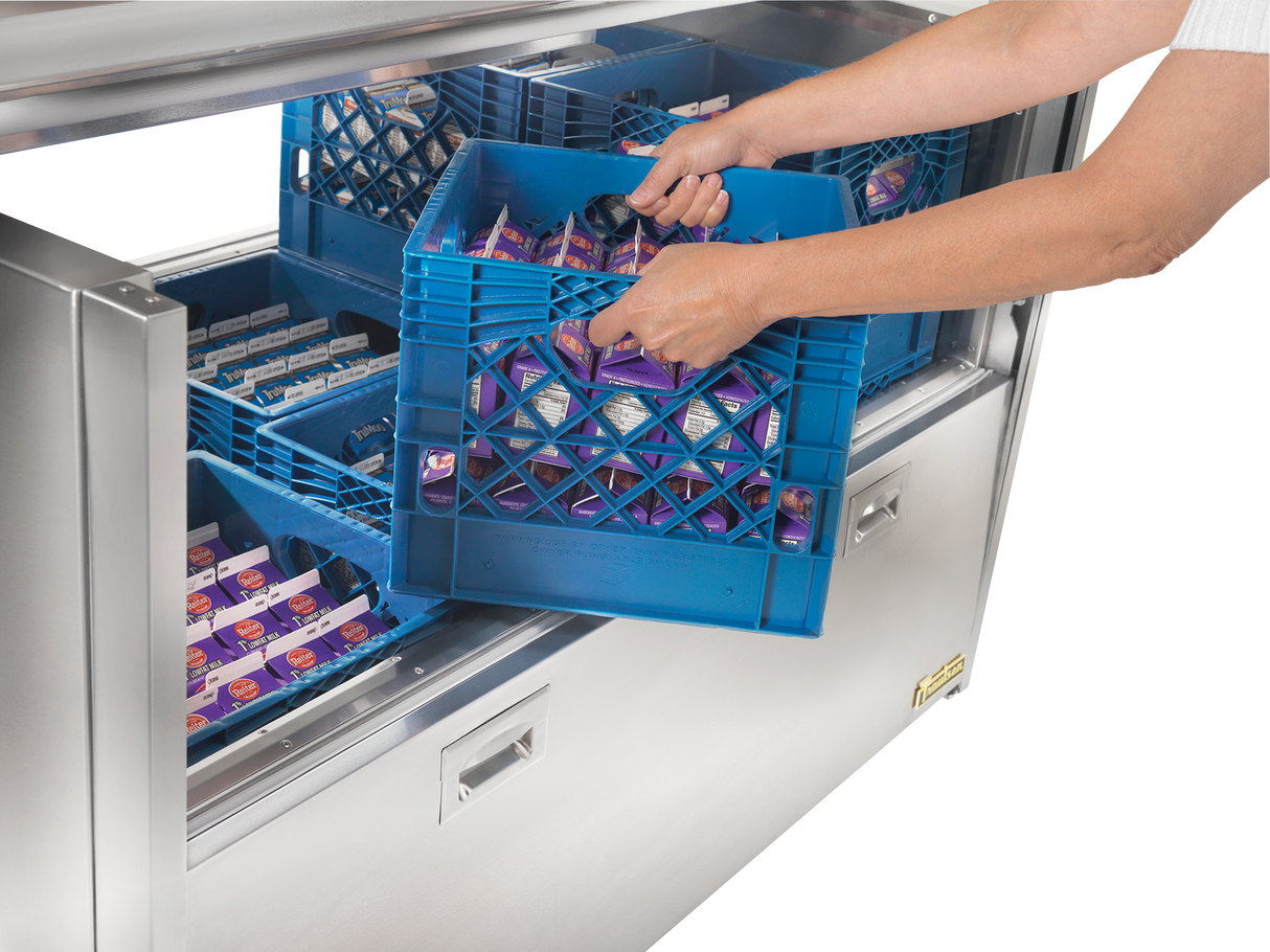 A person places a blue plastic crate filled with small cartons of milk into a stainless steel milk cooler drawer, which also contains other crates. Only the persons arms are visible.