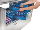 A person places a blue plastic crate filled with small cartons of milk into a stainless steel milk cooler drawer, which also contains other crates. Only the persons arms are visible.