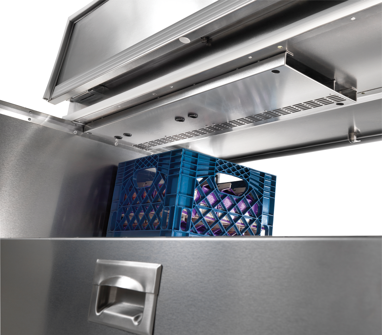 A blue plastic crate with purple bottles sits inside a Traulsen forced-air milk cooler, its stainless steel commercial unit open to reveal metal shelves and vents above the crate. The cooler features a brushed metal exterior and a silver handle in the foreground.