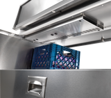 A blue plastic crate with purple bottles sits inside a Traulsen forced-air milk cooler, its stainless steel commercial unit open to reveal metal shelves and vents above the crate. The cooler features a brushed metal exterior and a silver handle in the foreground.