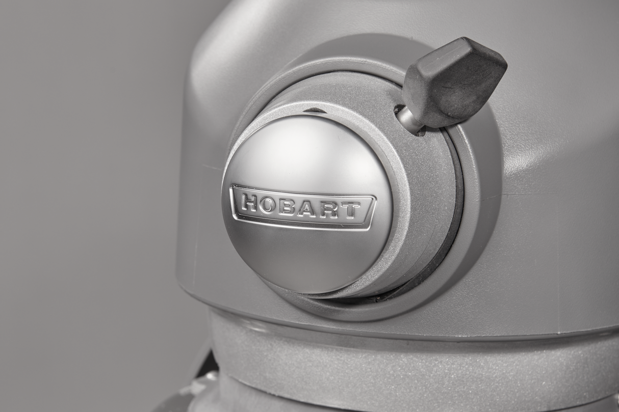 Close-up of a metallic Hobart planetary mixer head with a shiny round knob displaying the brand name “Hobart” in raised letters. A black lever extends from the side. The smooth, brushed finish stands out against a plain gray background.