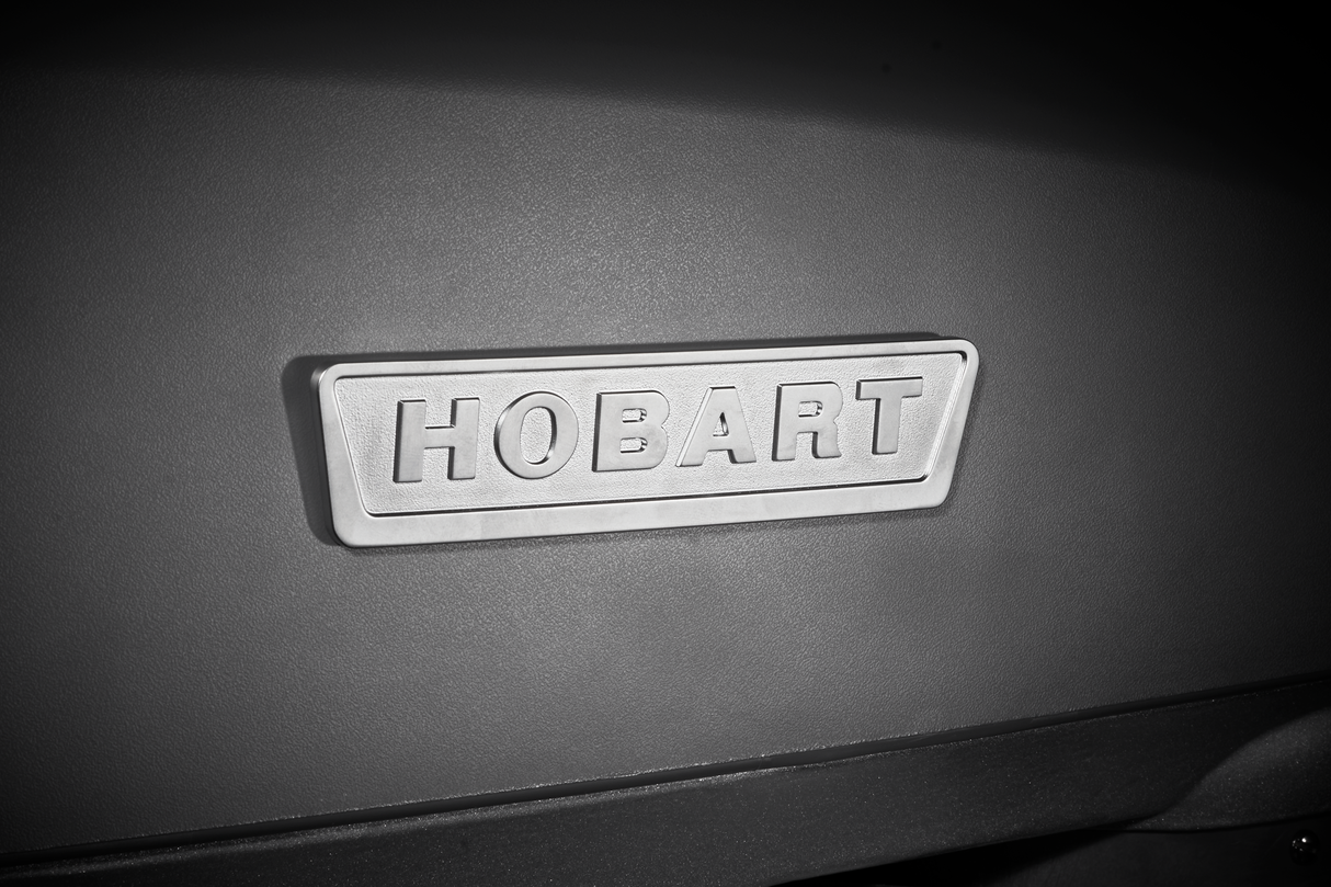 A close-up photo shows a metallic rectangular badge with the word “HOBART” embossed on it, attached to a smooth, dark gray surface of a 20 quart mixer. The lighting creates soft shadows and highlights, emphasizing the raised letters and texture.