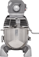 A large, industrial planetary mixer with a metallic finish is shown. It has a stainless steel bowl, wire safety guard, and a control knob on top. The sturdy base features a side lever. The background is plain white.