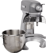 A large, industrial 20 quart planetary mixer in gray with a protective wire guard and metal mixing attachment. A shiny stainless steel mixing bowl with handles sits on the left, detached from the bench mixer, on a white background.