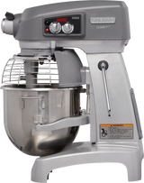 A large industrial planetary mixer with a metal bowl, safety guard, and control panel featuring two dials and a digital display. This 20 quart mixer is gray with a sturdy Hobart Legacy base and a yellow warning label on the lower side.