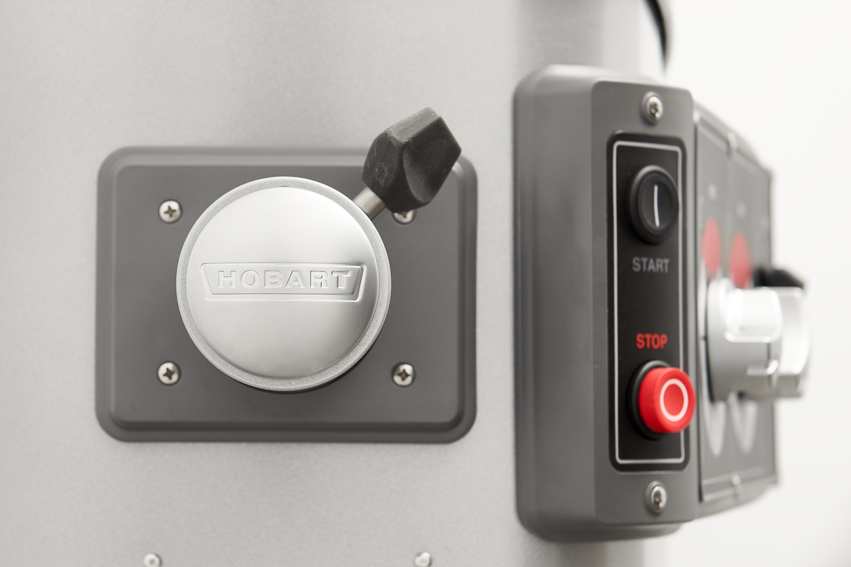 Close-up of a metallic Hobart mixer knob on the left, with a gray control panel featuring black, red, and white buttons labeled START and STOP on the right. The background is blurred, highlighting the Legacy Planetary Mixer’s details.