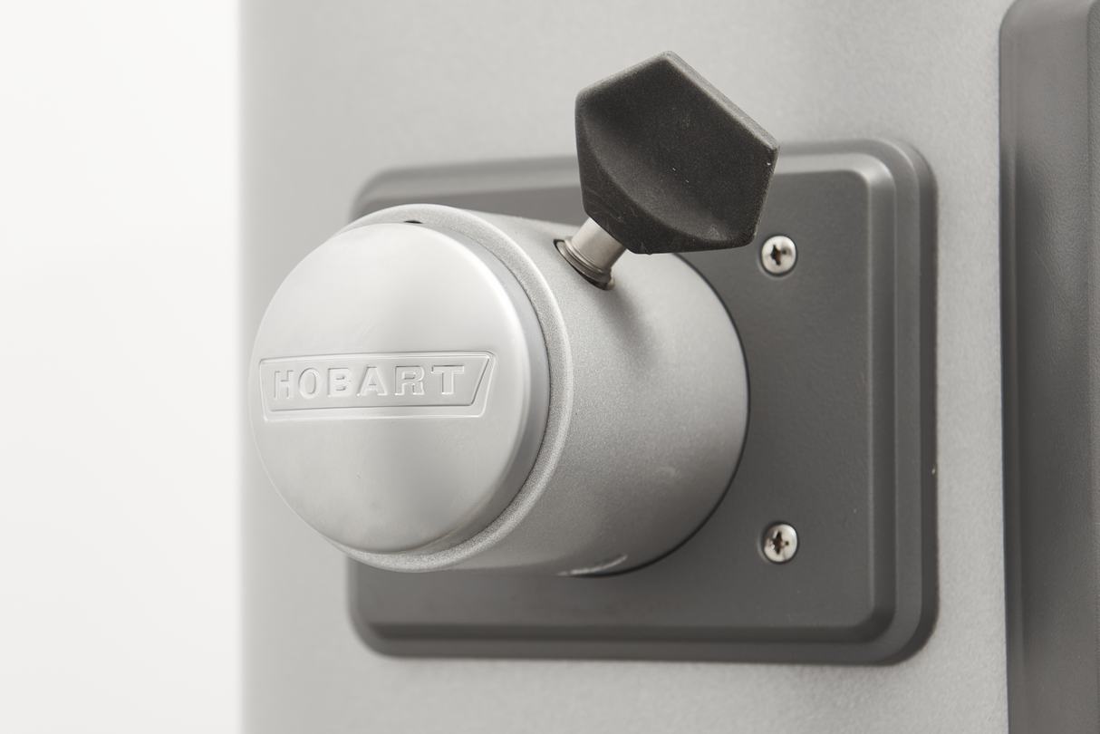 Close-up of a metallic knob with the brand name Hobart embossed on it, mounted on a gray panel with screws. A black, polygonal handle is attached above—a detail from a Hobart mixer, possibly part of a Legacy Planetary Mixer.