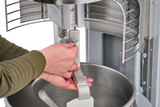 Close-up of hands adjusting the paddle on a Hobart HL600 Legacy Planetary Mixer, with a metal bowl below and safety guard above. The person in a green shirt works against a metallic mixer and background.
