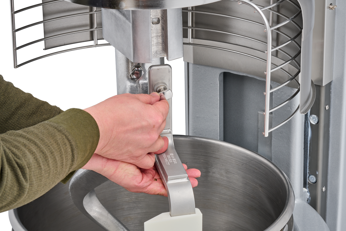 A person in a green sleeve uses both hands to tighten a metal attachment inside a 60 quart mixer, securing a paddle beater above a stainless steel mixing bowl. The Hobart HL600C correctional planetary mixer features a safety guard and metallic components.