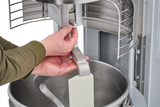 A person adjusts a metal attachment on a Hobart HL600 Legacy Planetary Mixer. One hand turns a knob, while the other supports a flat mixer blade inside the 60 quart stainless steel bowl, featuring safety guards and industrial design elements.
