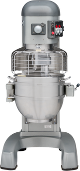 A large, industrial planetary mixer with a metallic body, protective wire guard, control panel with red and black buttons, and a thick mixing bowl sits on sturdy legs. A lever and other attachments are visible on the sides. The mixer is silver and gray.