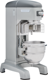 A large, industrial Legacy Planetary Mixer with a stainless steel bowl, safety guard, and control knobs on the top right. This Hobart 60 qt mixer is metallic gray, heavy-duty, and designed for commercial food prep on a sturdy base.