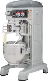 A large, industrial Hobart mixer with a metal mixing bowl and dough hook attachment. The commercial dough mixer is gray and silver, featuring control dials, safety instructions on the front panel, and a partial wire guard over the bowl.