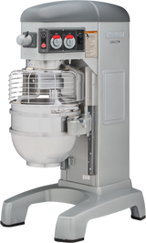 A large, industrial Hobart mixer with a stainless steel mixing bowl and protective wire guard. The metallic gray 60 qt mixer features control knobs, switches, and safety instructions on the front panel and stands on wide, sturdy legs.