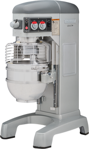 A large, industrial Hobart mixer with a stainless steel mixing bowl and protective wire guard. The metallic gray 60 qt mixer features control knobs, switches, and safety instructions on the front panel and stands on wide, sturdy legs.
