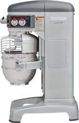 A large, industrial Hobart HL600C 60 quart mixer with a stainless steel bowl and protective wire guard. The Legacy Correctional Planetary Mixer features control knobs, a dial, warning labels, and a modern, utilitarian design on a sturdy base.