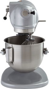 A silver 5 quart mixer with a large stainless steel mixing bowl and a wire whisk attachment. This planetary bench mixer features a rounded top with control levers, sits on a wide base, and secures the bowl in place with side latches.