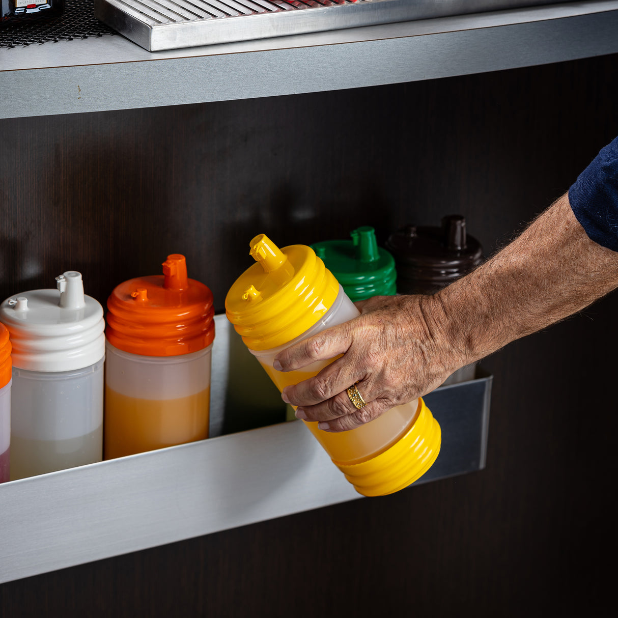 A hand grabs a yellow-lidded PourMaster Complete Unit squeeze bottle from a metal rack of five with color-coded caps—orange, white, green, brown—each filled with various liquids, against a dark background. All bottles are BPA free.