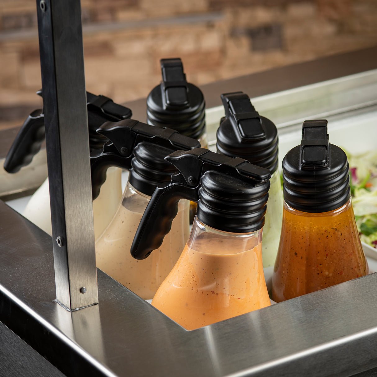Four polycarbonate salad dressing dispensers with black handles are arranged in a row at a salad bar. Each dishwasher safe bottle contains a different colored dressing—white, orange, and reddish-brown. A portion of mixed salad is visible to the right.