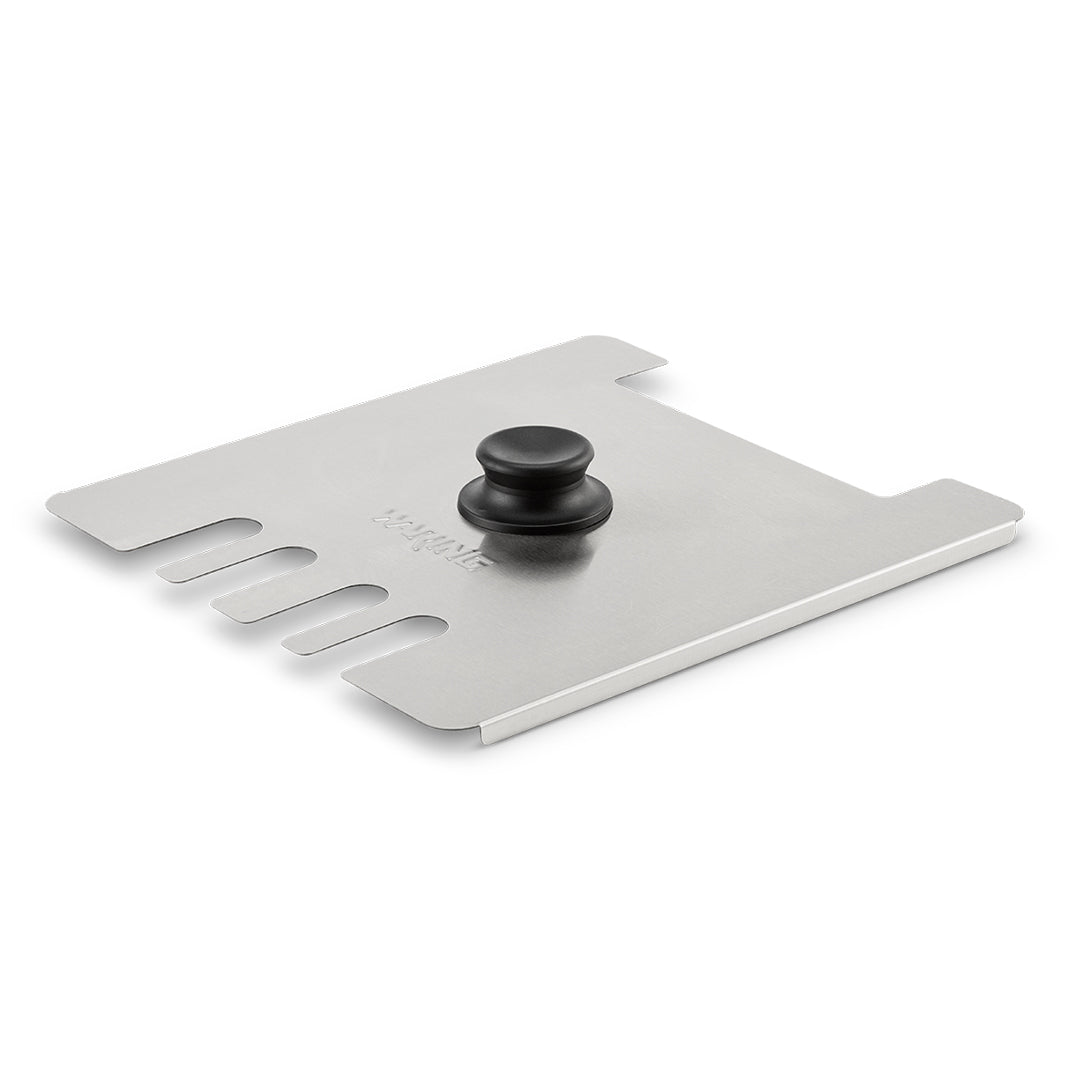 A rectangular stainless steel grill scraper with a black knob handle in the center, similar to a Waring deep fryer accessory. The left edge features five curved notches for cleaning grates; the opposite side has two square cutouts and straight edges. “Sur La Table” is visible.