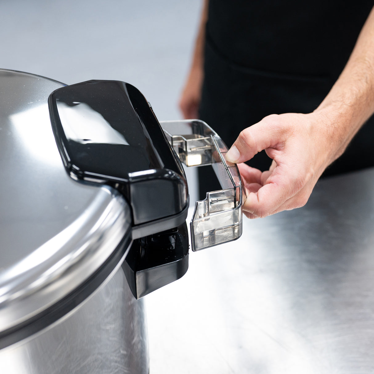A person’s hand is turning the clear plastic handle on the lid of a large stainless steel rice cooker, likely a commercial rice cooker with 20 cup capacity, on a shiny metal countertop. The background and the person’s torso are out of focus.
