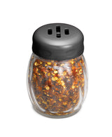 A clear glass shaker filled with red pepper flakes, featuring a BPA free black plastic lid with three oval-shaped holes for dispensing. The shaker is photographed against a plain white background.