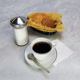 A cup of black coffee on a saucer with a spoon and napkin sits beside a glass sugar dispenser with a stainless steel pourer. Behind them, a basket lined with yellow cloth holds assorted cookies and pastries on the light marble surface.