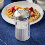 A stainless steel shaker filled with powdered sugar sits on a blue cloth. In the background, a plate holds a waffle topped with chopped strawberries, creating a cozy breakfast scene. The shaker is also dishwasher safe for easy cleaning.