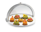 A white plate with eight colorful, decorated miniature pastries is displayed under a half-opened polycarbonate dome cover. The pastries are round, topped with chocolate shapes, fruit pieces, and decorative accents in green, orange, and red.