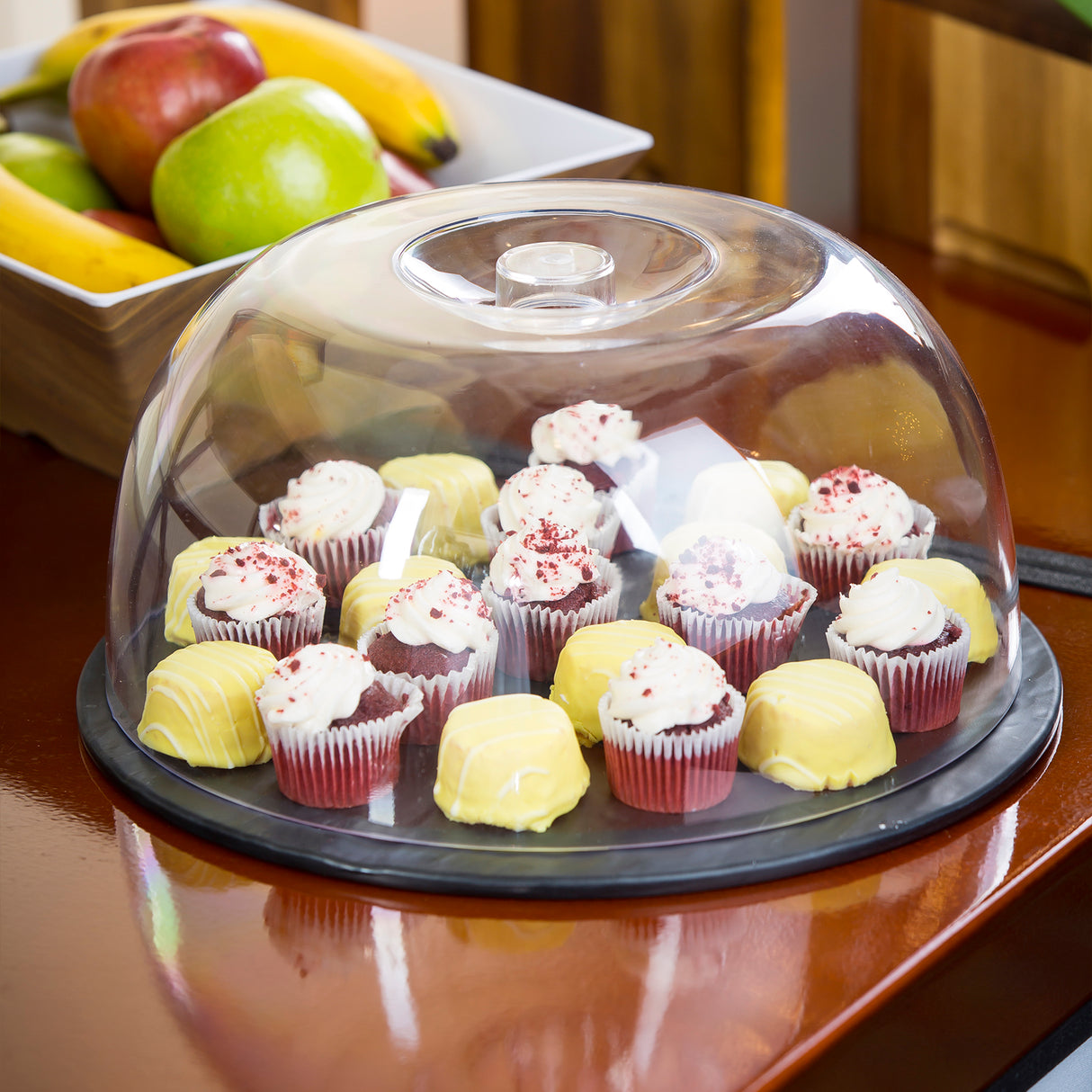 A platter of assorted mini desserts rests under a 12" dia Cake Dome, keeping red velvet cupcakes and yellow-frosted treats fresh. The clear cover is dishwasher safe, while a bowl of bananas and apples adds color in the background.