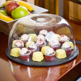 A platter of assorted mini desserts rests under a 12" dia Cake Dome, keeping red velvet cupcakes and yellow-frosted treats fresh. The clear cover is dishwasher safe, while a bowl of bananas and apples adds color in the background.