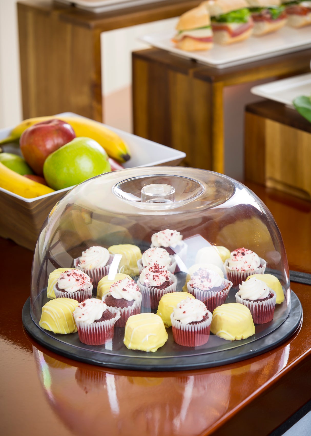 A platter of assorted mini cupcakes and desserts is covered by a clear TableCraft cake dome and sits on a wooden table. In the background, a white bowl of apples and bananas, and a tray with a sandwich on a raised wooden stand add charm.