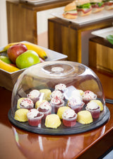 A platter of assorted mini cupcakes and desserts is covered by a clear TableCraft cake dome and sits on a wooden table. In the background, a white bowl of apples and bananas, and a tray with a sandwich on a raised wooden stand add charm.