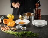 A chef ladles orange soup into a white bowl. On the dark countertop are squash, carrots, ginger, diced pumpkin, a pot of soup, stacked plates, a TableCraft pepper mill, and small bowls of seeds and herbs. Fresh carrot tops are in the foreground.