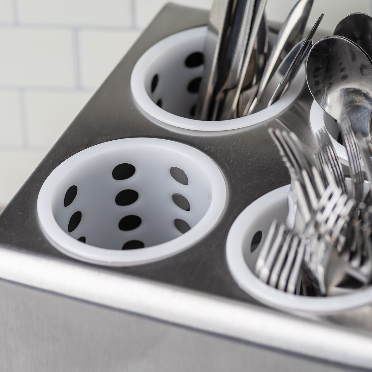 A close-up of a stainless steel cutlery holder with white, perforated polypropylene flatware cylinders. Some cups hold metal forks and utensils, while others are empty. The background shows a white tiled wall with a subtle grid pattern.