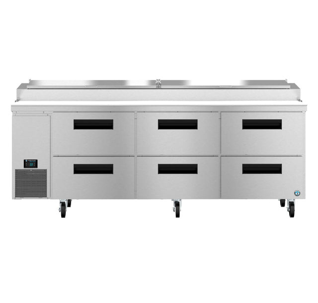 A large commercial stainless steel pizza prep table with six black-handled refrigerated drawers, a digital control panel on the left, and four caster wheels. The top features a covered compartment for food storage or ingredients.