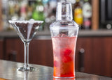A clear 20 oz cocktail shaker with a built-in strainer, filled with ice and a red drink, sits on a bar counter. Next to it is an empty martini glass with a salt-rimmed edge, against a backdrop of blurred bottles on shelves.