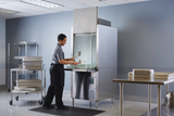 A person in a gray shirt and black apron loads metal utensils into an Advansys Hobart commercial utensil washer in a clean, industrial kitchen. Nearby, stainless steel tables and carts with trays stand on the gray floor beside light blue walls.